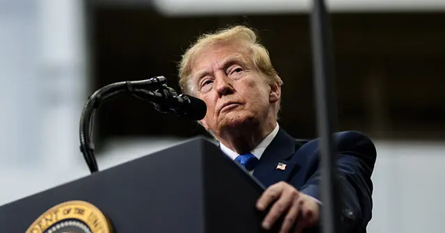 The image shows a person standing at a podium with a microphone, wearing a suit and tie, and appears to be addressing an audience.