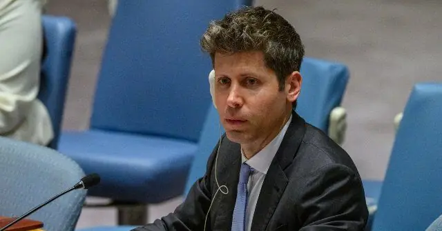 The image shows a man in a suit seated at a table with a microphone, likely participating in a formal meeting or conference.