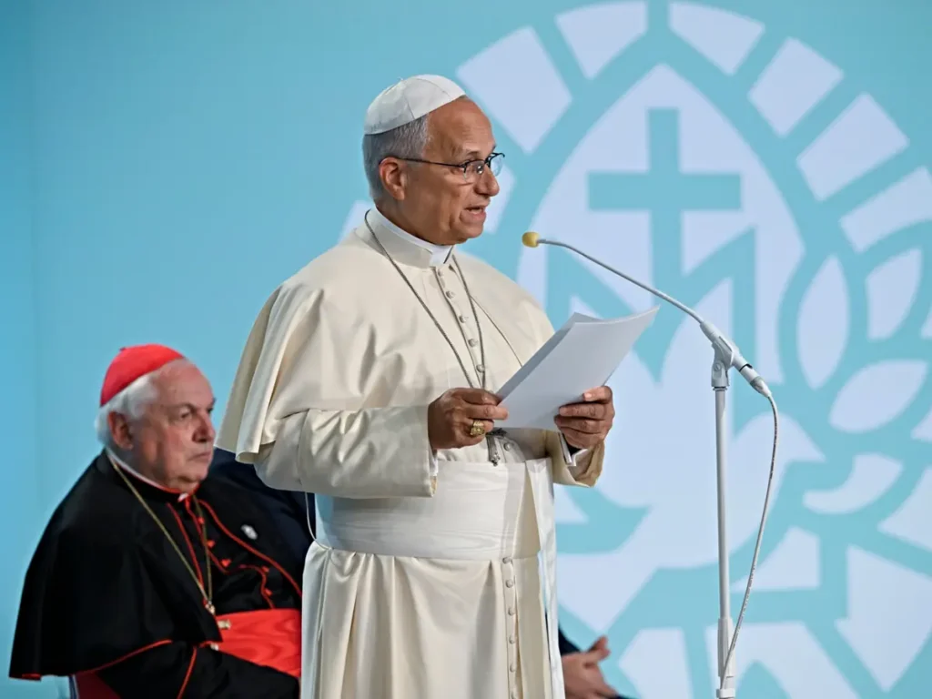 Pope Francis delivers a speech while holding a document, with a cardinal seated in the background.