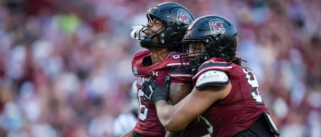 Two football players in maroon uniforms celebrate on the field during a game.