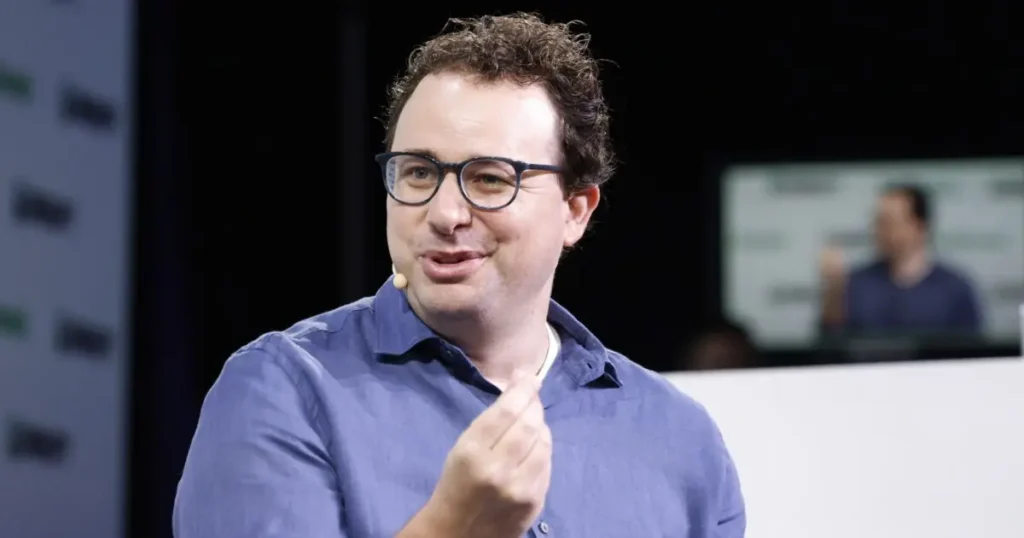 The image shows a man wearing glasses and a blue shirt, gesturing with his hand while speaking, likely during a presentation or speech.