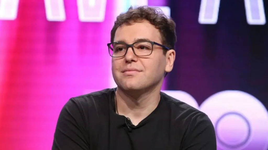 The image shows a person wearing glasses and a black shirt, sitting in front of a colorful background with illuminated shapes.