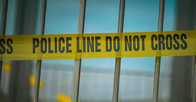 The image shows a "POLICE LINE DO NOT CROSS" tape stretched across a metal fence, indicating a restricted area.