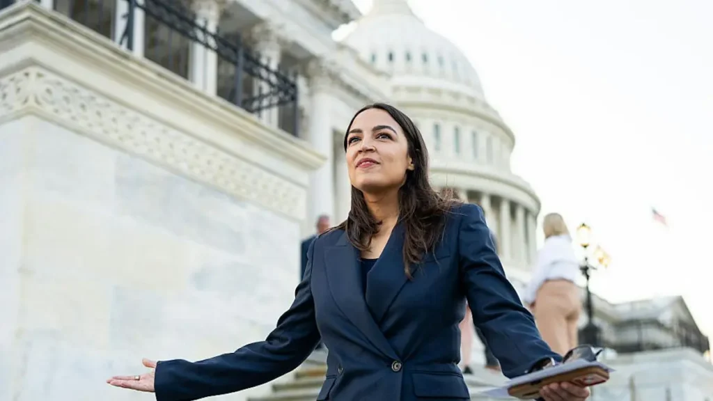 A woman in a suit stands in front of the U.S. Capitol, holding a folder and gesturing with her hands.