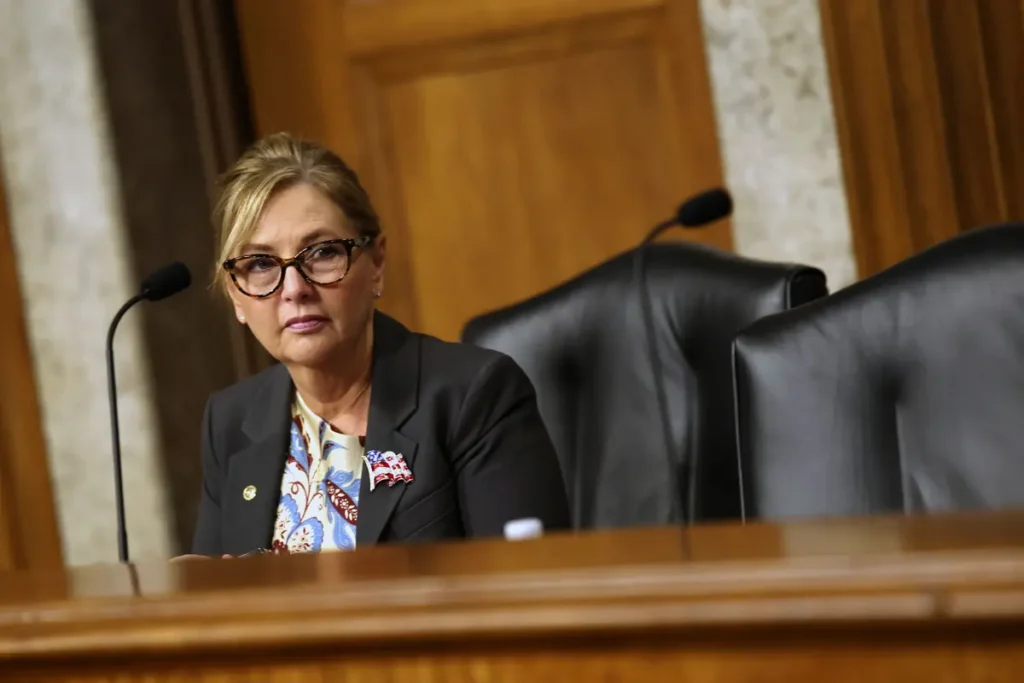 A woman in formal attire is seated at a desk with microphones, suggesting she is participating in a formal or official proceeding.