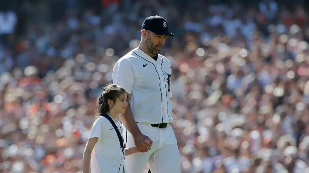 A baseball player and a young girl walk together on the field during a game, with a crowd in the background.