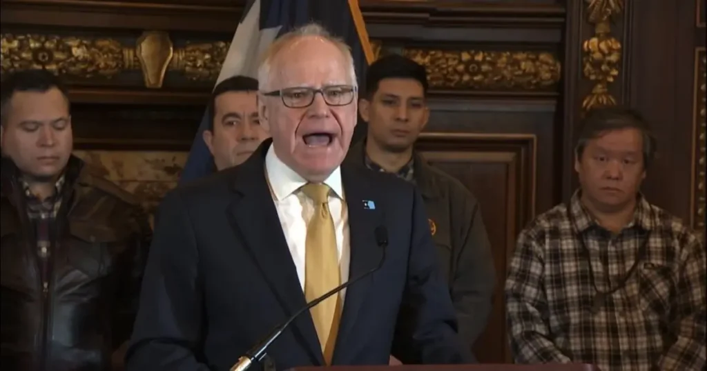 A man in a suit and tie is speaking at a podium in front of a group of people and a flag.