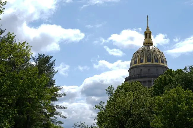 The image shows the golden dome of the West Virginia State Capitol building, surrounded by trees under a partly cloudy sky.