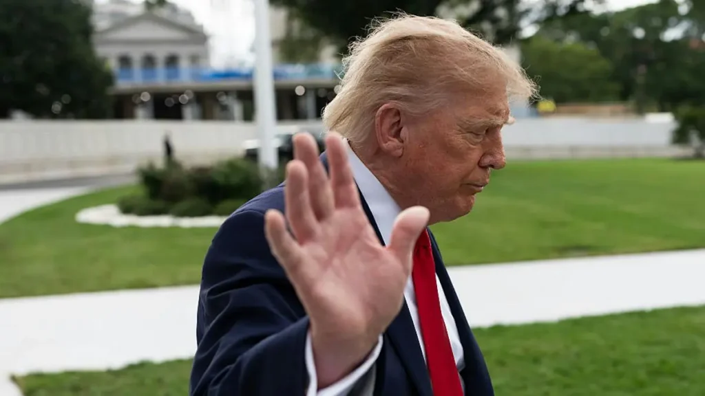 The image shows a man in a suit and tie waving with his right hand, with a building and greenery in the background.
