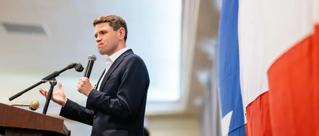 A man in a suit is giving a speech at a podium with a microphone, with a flag in the background.