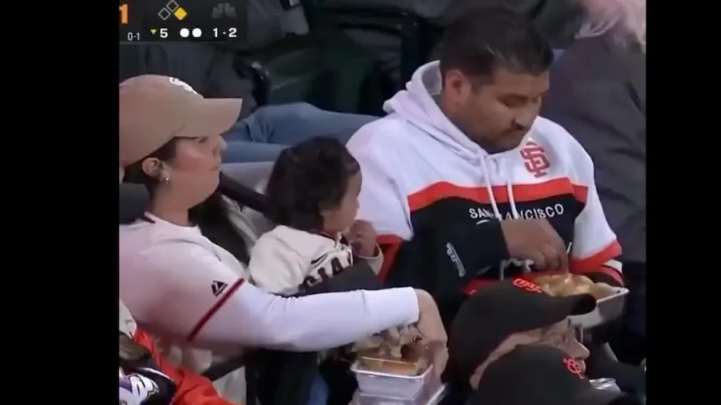 A family enjoys a meal together at a baseball game, with the father wearing a San Francisco Giants hoodie.