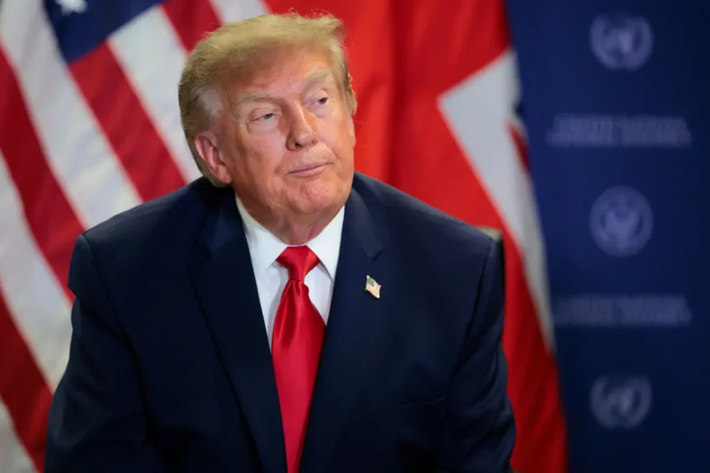 The image shows a person in a dark suit and red tie, seated in front of an American flag and a backdrop with the G20 logo.