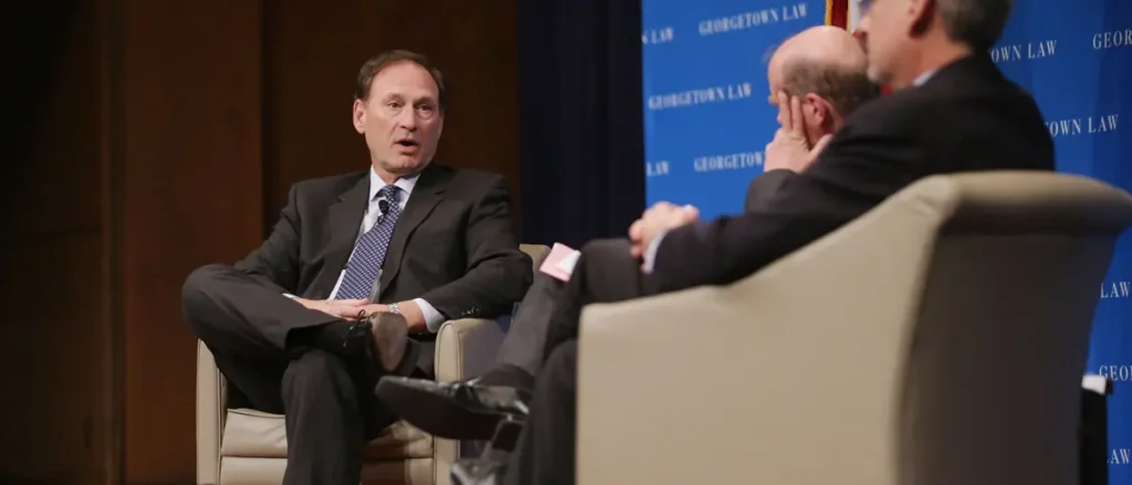 The image shows a panel discussion at Georgetown Law, with a man in a suit speaking while another listens attentively.