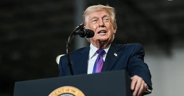 The image shows a person speaking at a podium with a microphone, dressed in a suit and tie, and wearing a small American flag pin on the lapel.