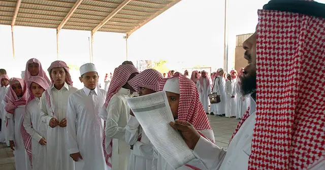 A group of young men in traditional attire are gathered under a covered structure, with one individual holding a document.