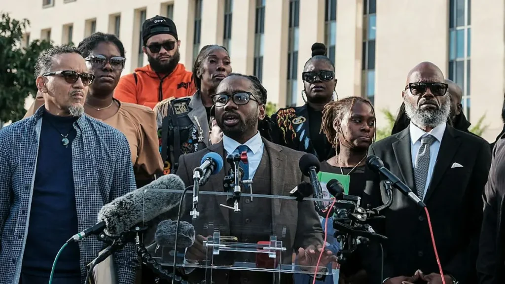A group of individuals stands behind microphones at a press conference, with one person speaking at a podium.