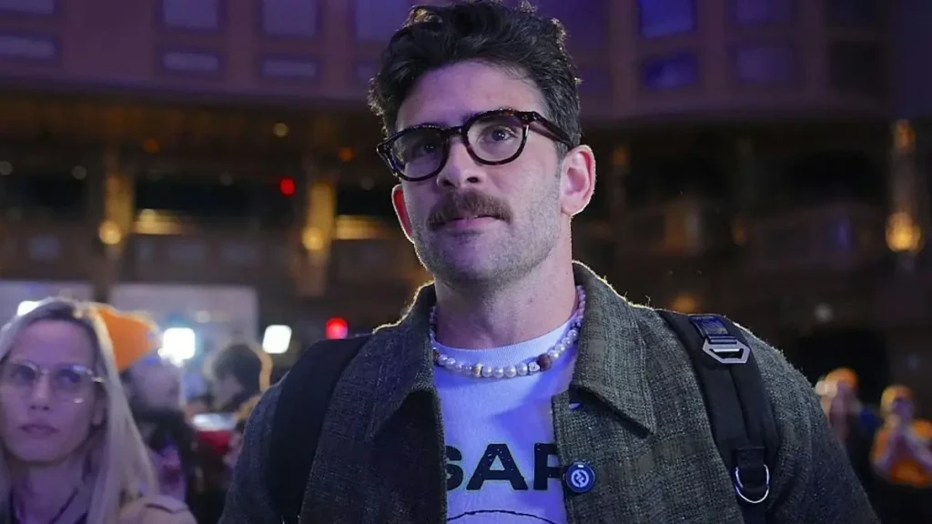 The image shows a man wearing glasses, a pearl necklace, and a "SAP" t-shirt, standing in a crowded urban setting at night.