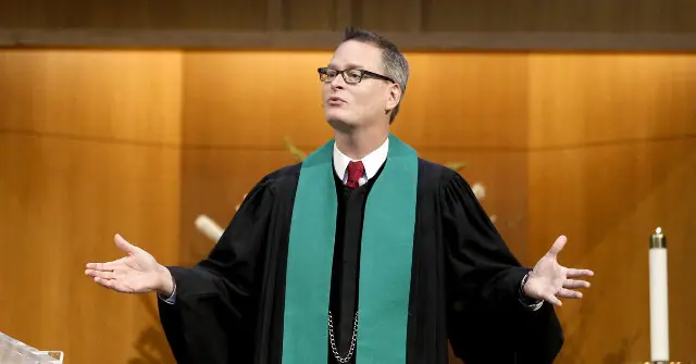 A person in a black robe and green stole stands with arms outstretched in a church setting.