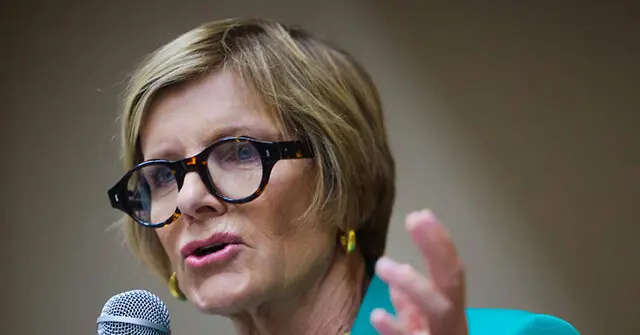 A woman wearing glasses and a turquoise top is speaking into a microphone, gesturing with her hand.