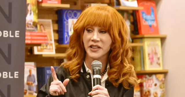 A woman with red hair is speaking into a microphone in front of a bookshelf.