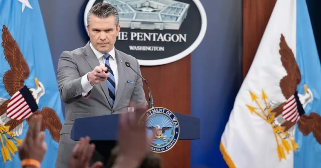 A man in a suit stands at a podium with the Pentagon logo and Department of War seal, addressing a press conference.