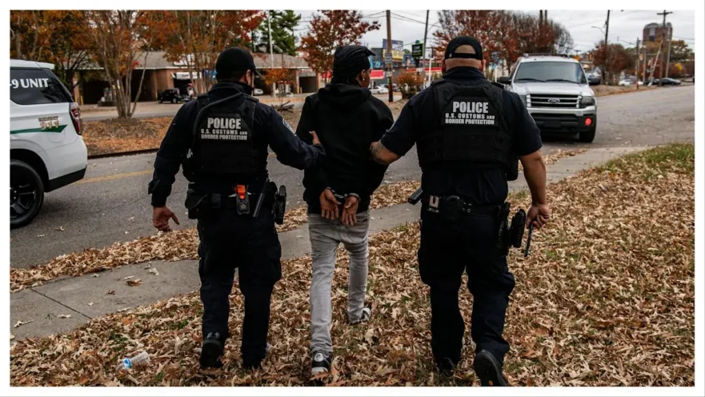 Two U.S. Customs and Border Protection officers escort a handcuffed individual along a sidewalk.