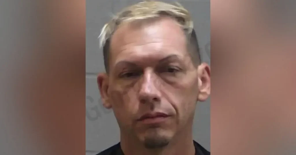 The image appears to be a mugshot of a man with short, light-colored hair, wearing a dark shirt.