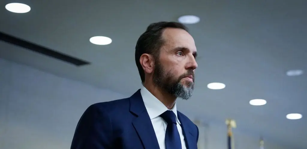 The image shows a man in a suit and tie, looking off to the side in a formal setting.