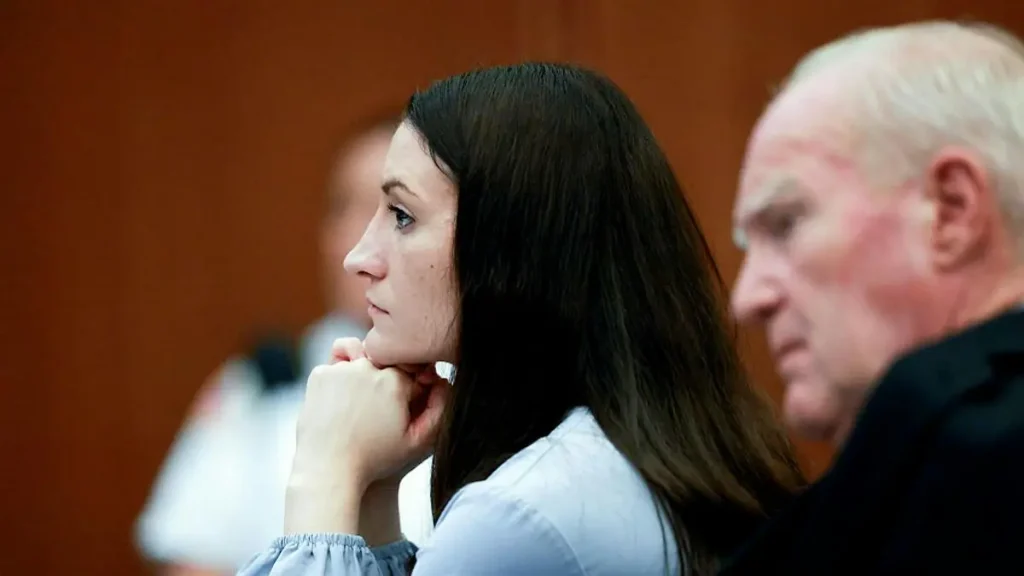 The image shows a woman sitting in a courtroom with her hand resting on her chin, appearing contemplative or concerned.