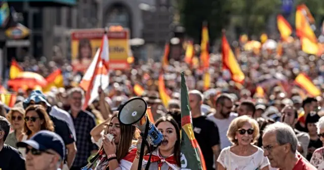 The image depicts a large crowd gathered outdoors, holding flags and participating in a public demonstration or rally.