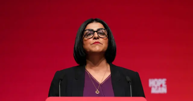 A woman stands at a podium in front of a red background, delivering a speech.