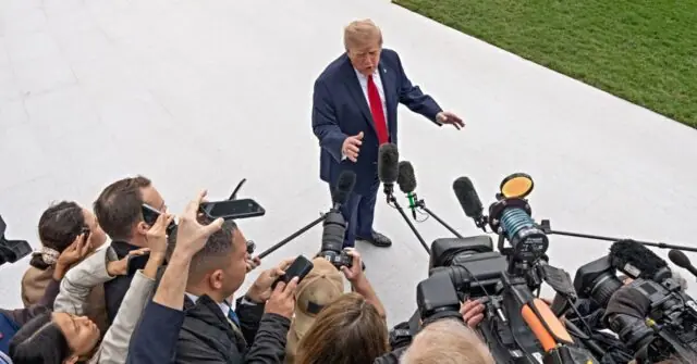 The image shows a public figure addressing a group of reporters and photographers, surrounded by microphones and cameras.