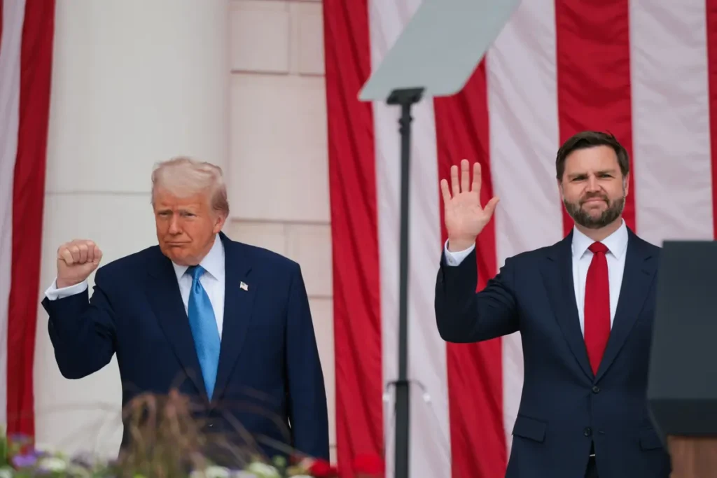The image shows two men in suits standing in front of American flags, with one man raising his hand in a wave and the other making a fist gesture.