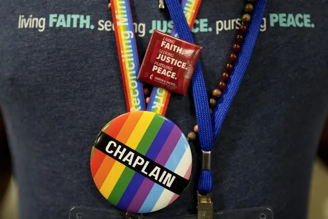 The image shows a lanyard with a badge that reads "CHAPLAIN" and a rainbow-colored pin, symbolizing LGBTQ+ pride, attached to it.