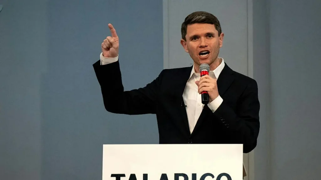 A man in a suit is giving a speech at a podium with a microphone, gesturing with his right hand.
