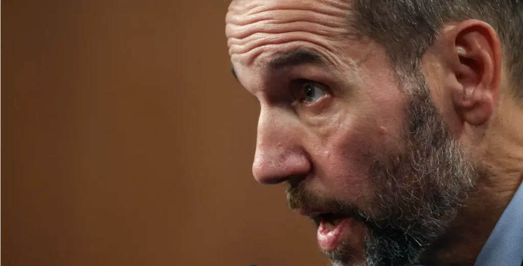 The image shows a close-up of a man with a beard and a light blue shirt, speaking with his mouth slightly open.