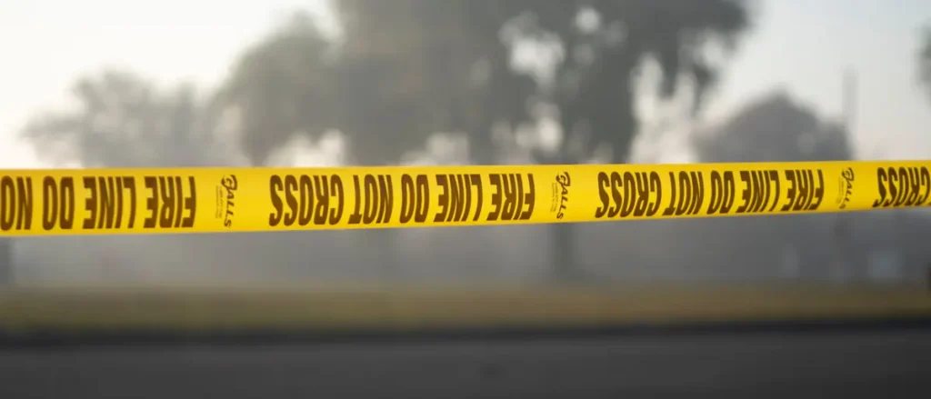 A "Fire Line Do Not Cross" yellow caution tape is stretched across a scene with smoke in the background, indicating a fire or emergency situation.