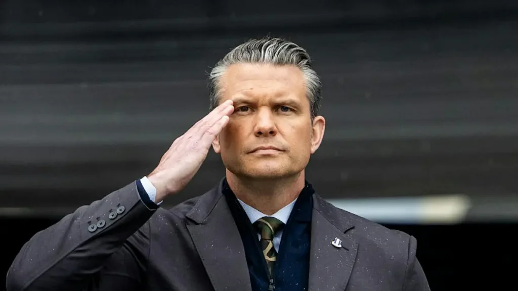 A man in a suit salutes with his right hand, standing against a dark background.