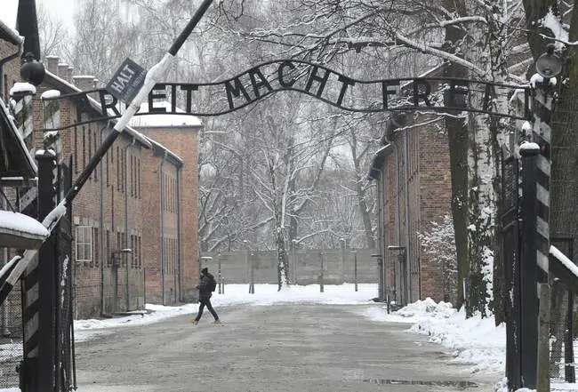 The image shows the entrance gate of Auschwitz-Birkenau, with the sign "Arbeit macht frei" (Work sets you free) partially covered in snow, symbolizing the historical and somber nature of the site.