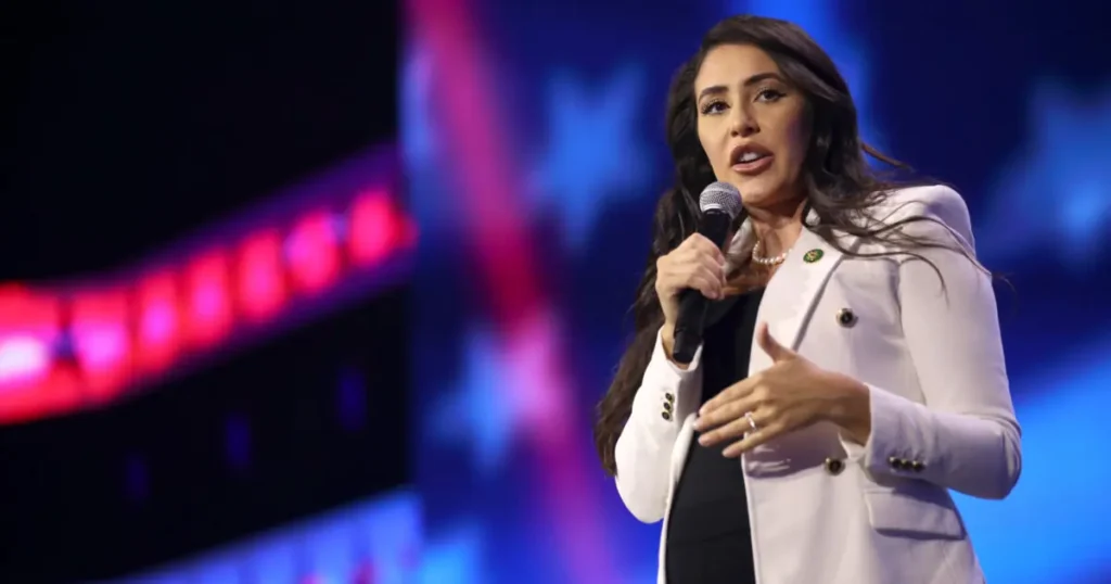 A woman in a white blazer is speaking into a microphone on a stage with colorful lighting in the background.