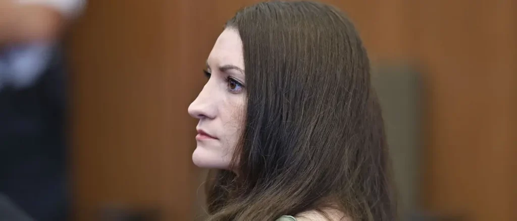 The image shows a person with long brown hair seated in what appears to be a formal setting, possibly a courtroom or a professional environment.