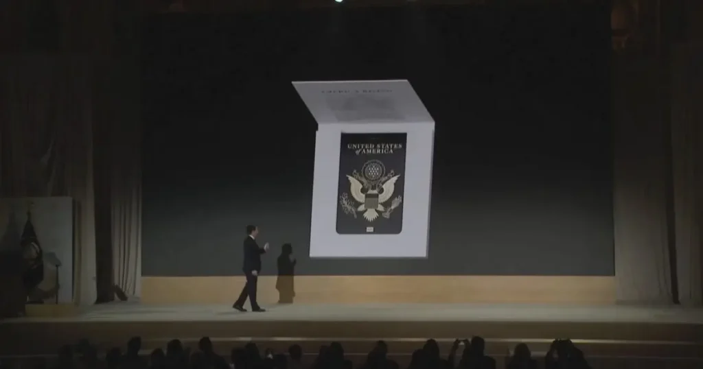 A man in a suit stands on stage in front of a large screen displaying the seal of the United States of America.
