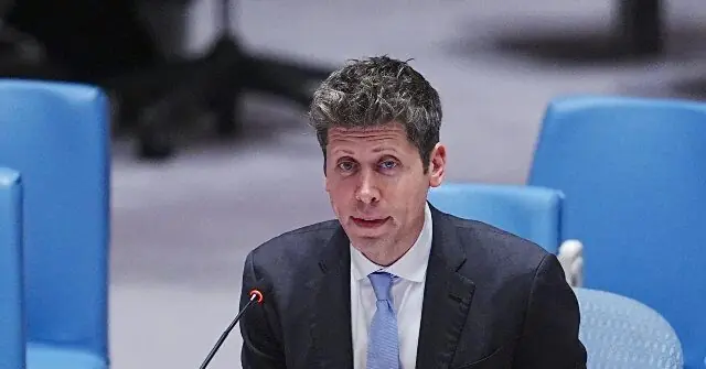 The image shows a man in a suit speaking at a formal event, with blue chairs and a blurred background suggesting a conference or meeting setting.