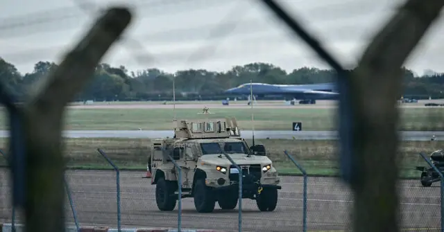 A military vehicle is seen on a runway with a large aircraft in the background, viewed through a fence.