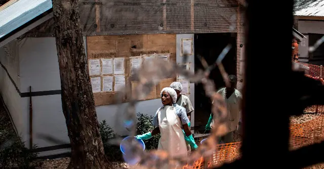 The photo shows a group of individuals in protective clothing and gloves working outside a building with a bulletin board, possibly in a rural or agricultural setting.