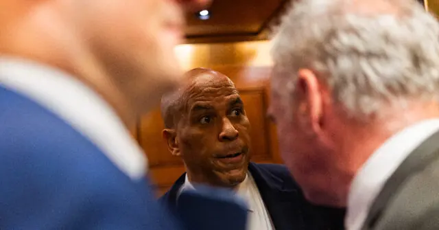 The image shows a man in a suit engaged in a conversation with another individual, with a third person partially visible in the foreground.