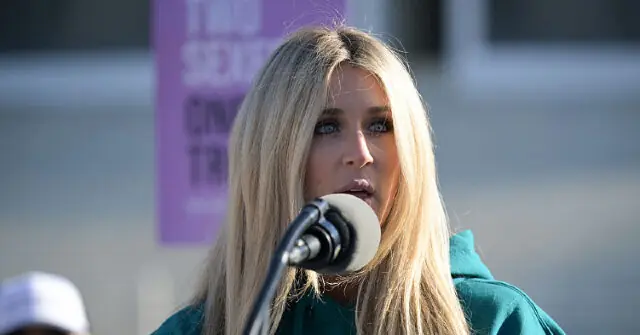 A person with long blonde hair is speaking into a microphone at an outdoor event.