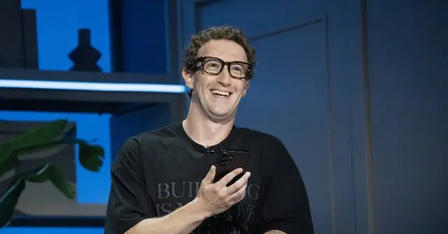 The image shows a person smiling while holding a smartphone, wearing a black t-shirt with text on it, in a setting with a blue background and some decorative elements.