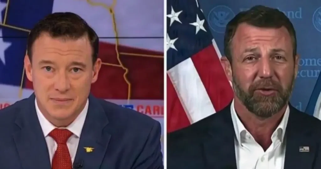 The image shows two individuals in formal attire, one wearing a suit with a red tie and the other in a suit with a white shirt, both set against a backdrop featuring an American flag and political imagery.