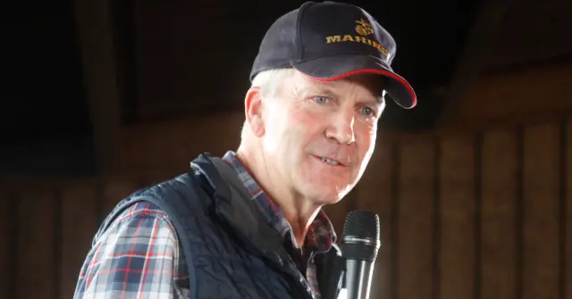 The image shows a man wearing a Marine Corps baseball cap and a plaid shirt, speaking into a microphone in what appears to be a formal or public speaking setting.